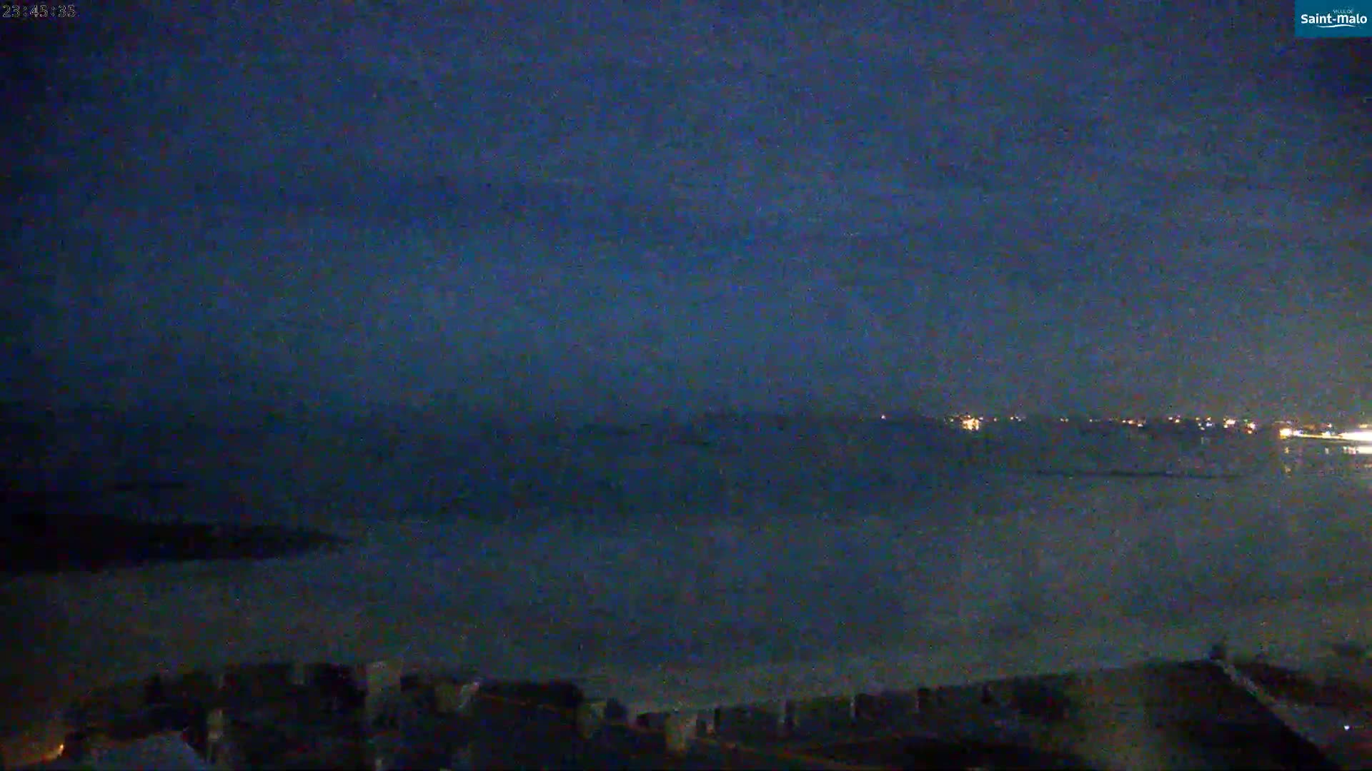 A dark, nighttime view shows a calm body of water with a distant, dimly lit shoreline under a mostly clear, dark blue sky.