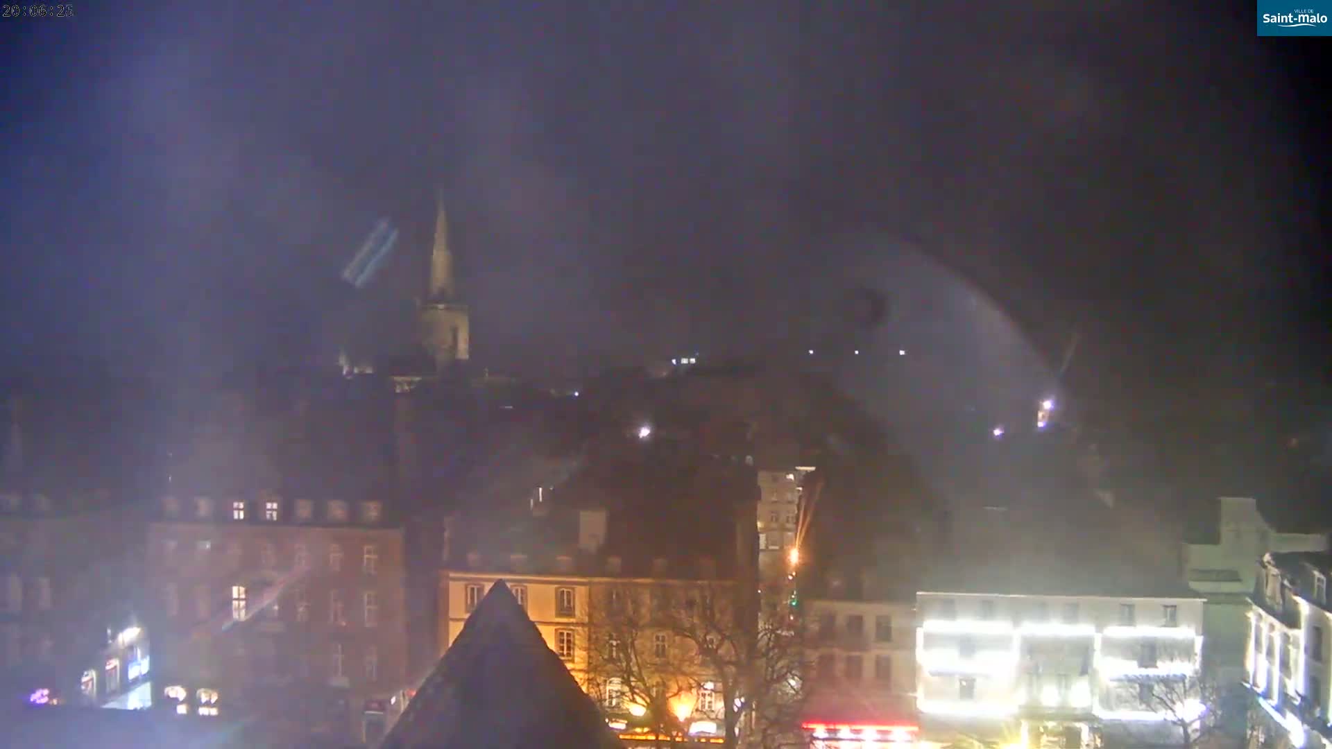 A nighttime view of a town, possibly in Europe, with a tall church spire visible in the distance under a hazy, dark sky.