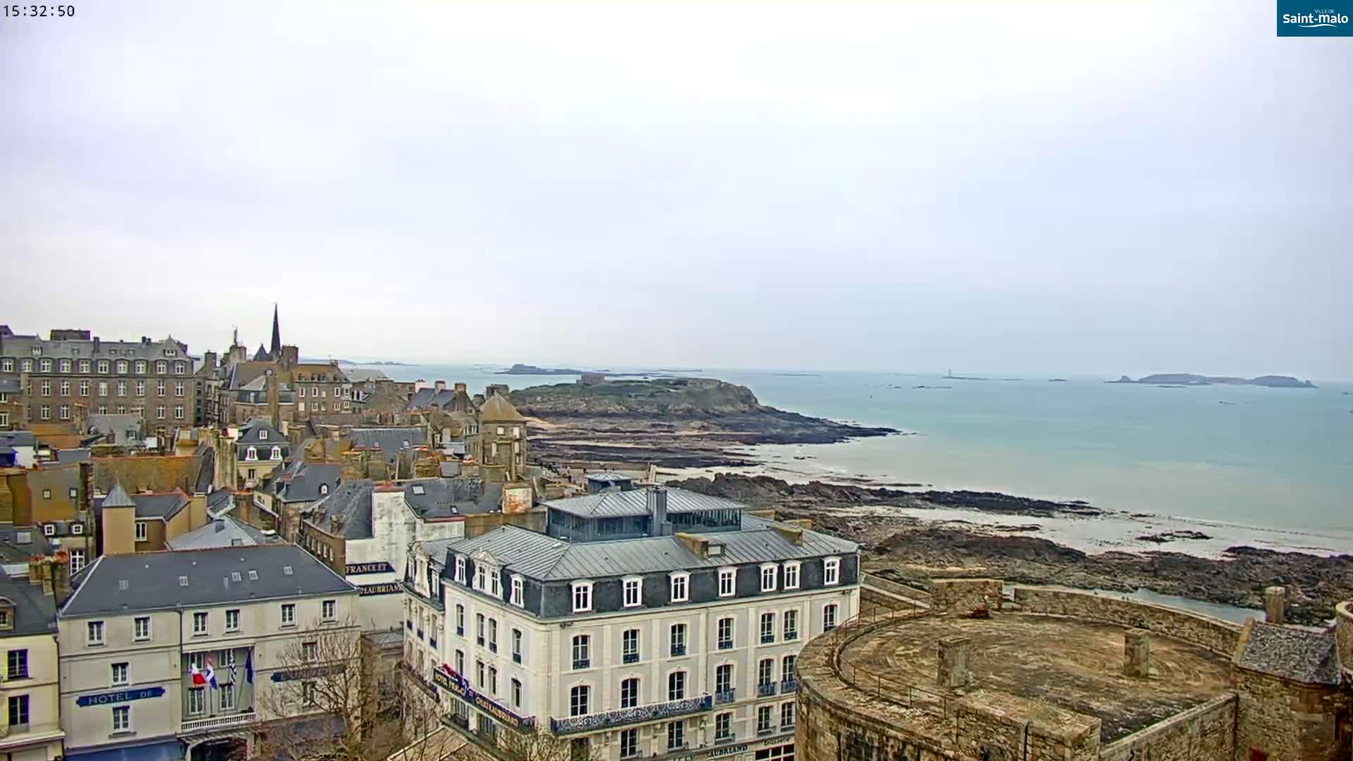 A coastal town with stone buildings overlooks a rocky shoreline and calm ocean under an overcast sky.
