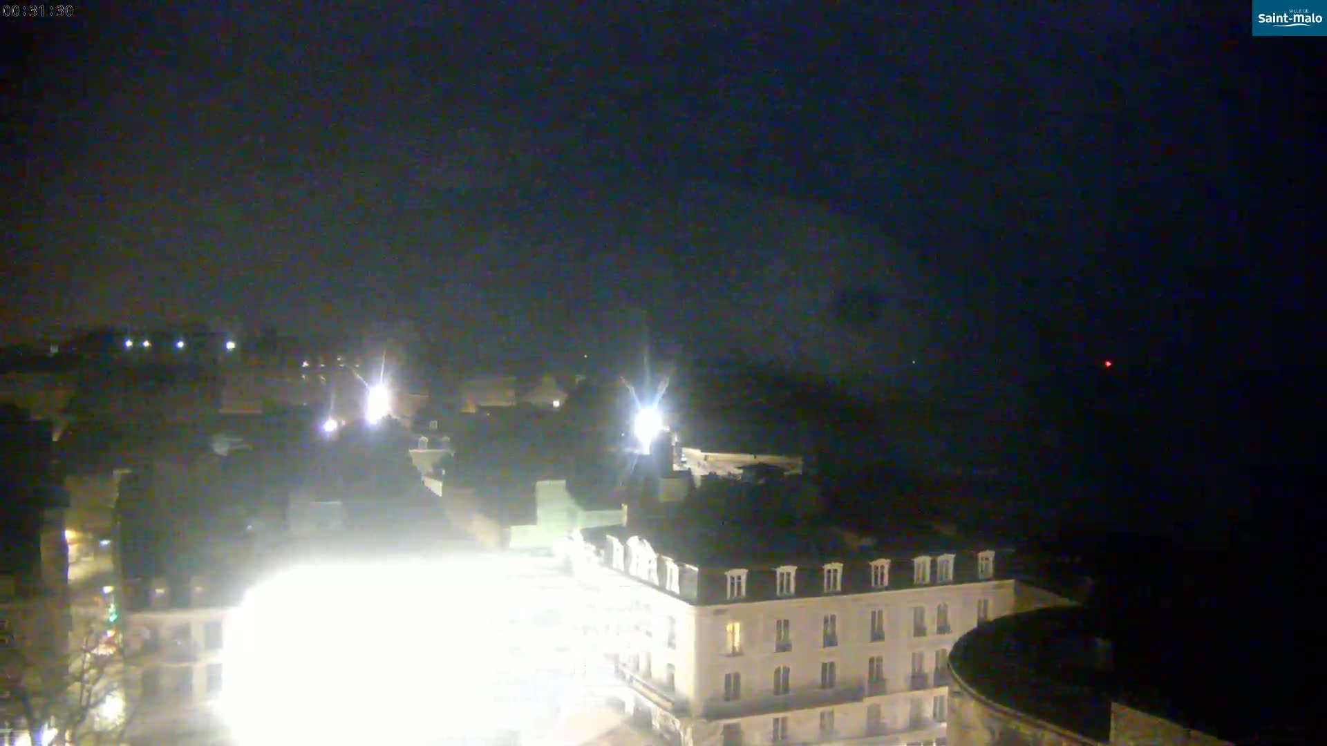 A nighttime, high-angle view shows a city square with a multi-story building and surrounding buildings illuminated by bright lights under a dark, clear sky.