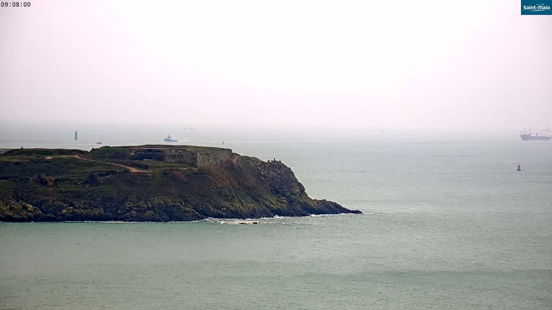 A rocky, green peninsula juts into a calm, hazy sea where several ships are visible in the distance.