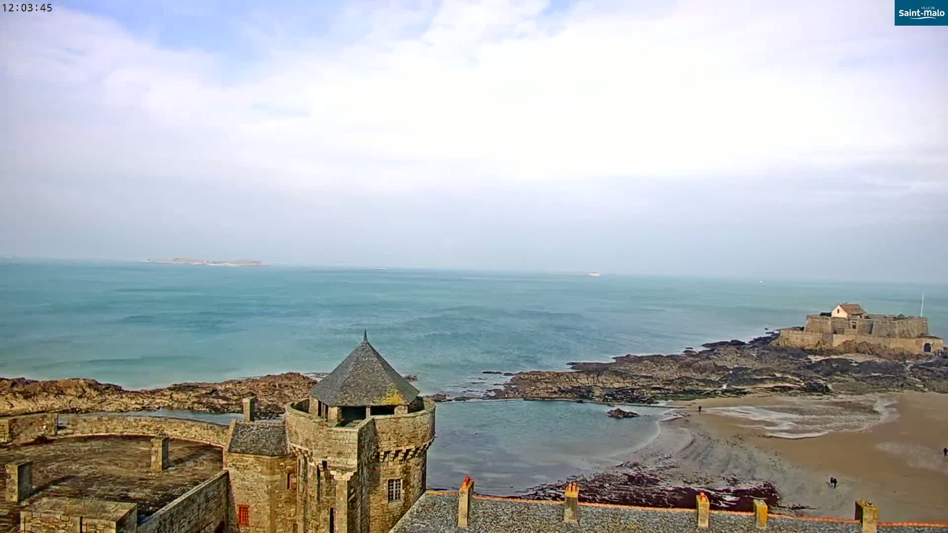 A stone castle overlooks a calm, teal ocean with a small island in the distance under a mostly cloudy sky.