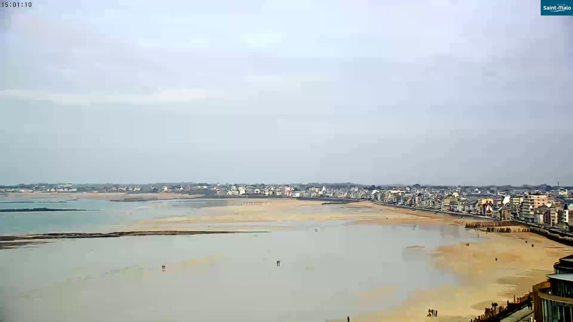 A coastal town is visible under an overcast sky, with a wide sandy beach and calm, shallow water extending to a body of darker water beyond.