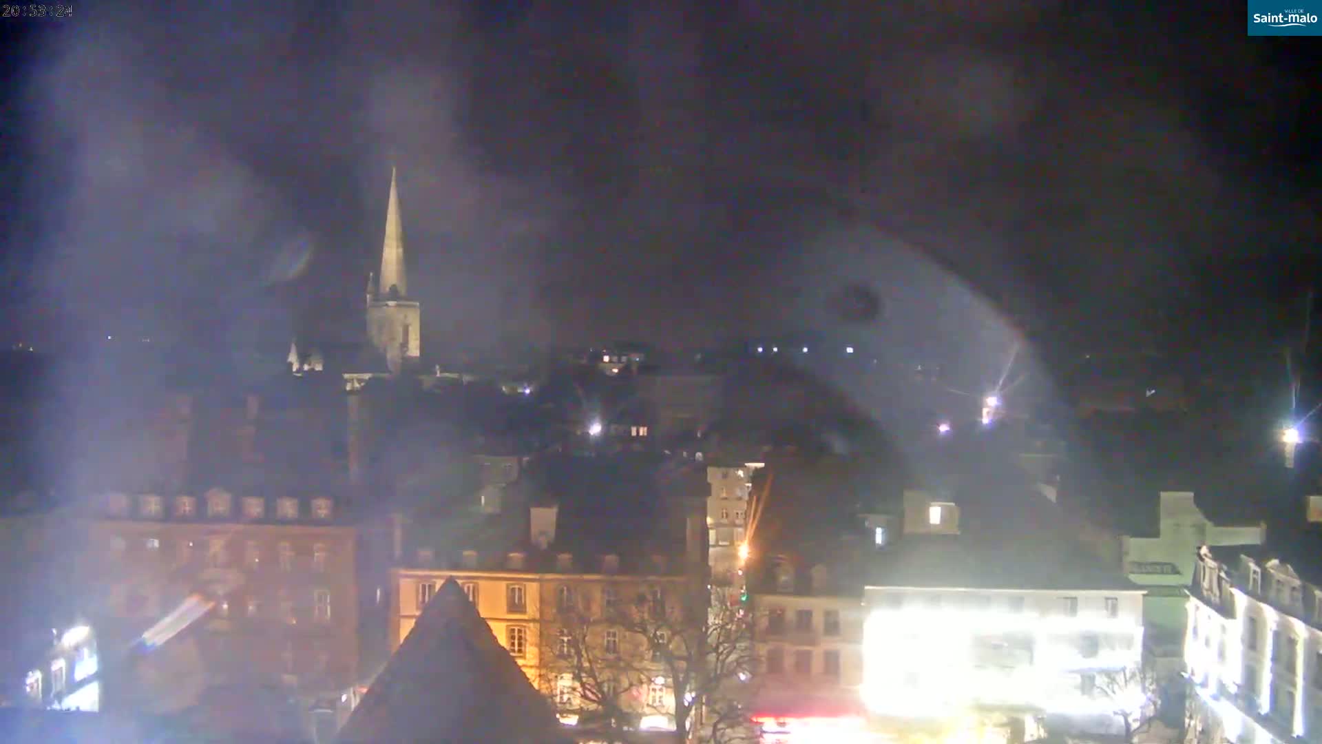A nighttime view of a city with a tall church spire in the background, under hazy or foggy conditions.
