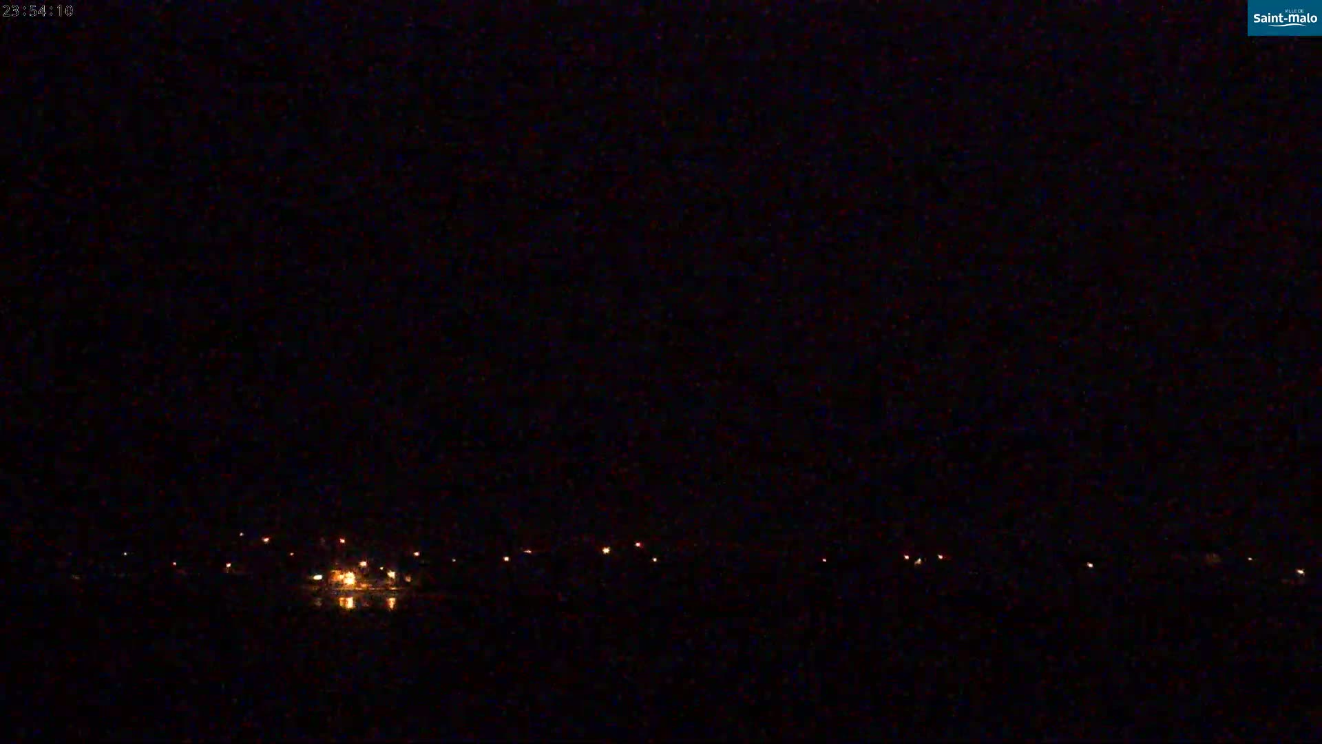 A dark nighttime scene shows a line of distant lights reflecting on dark water under a clear, black sky.