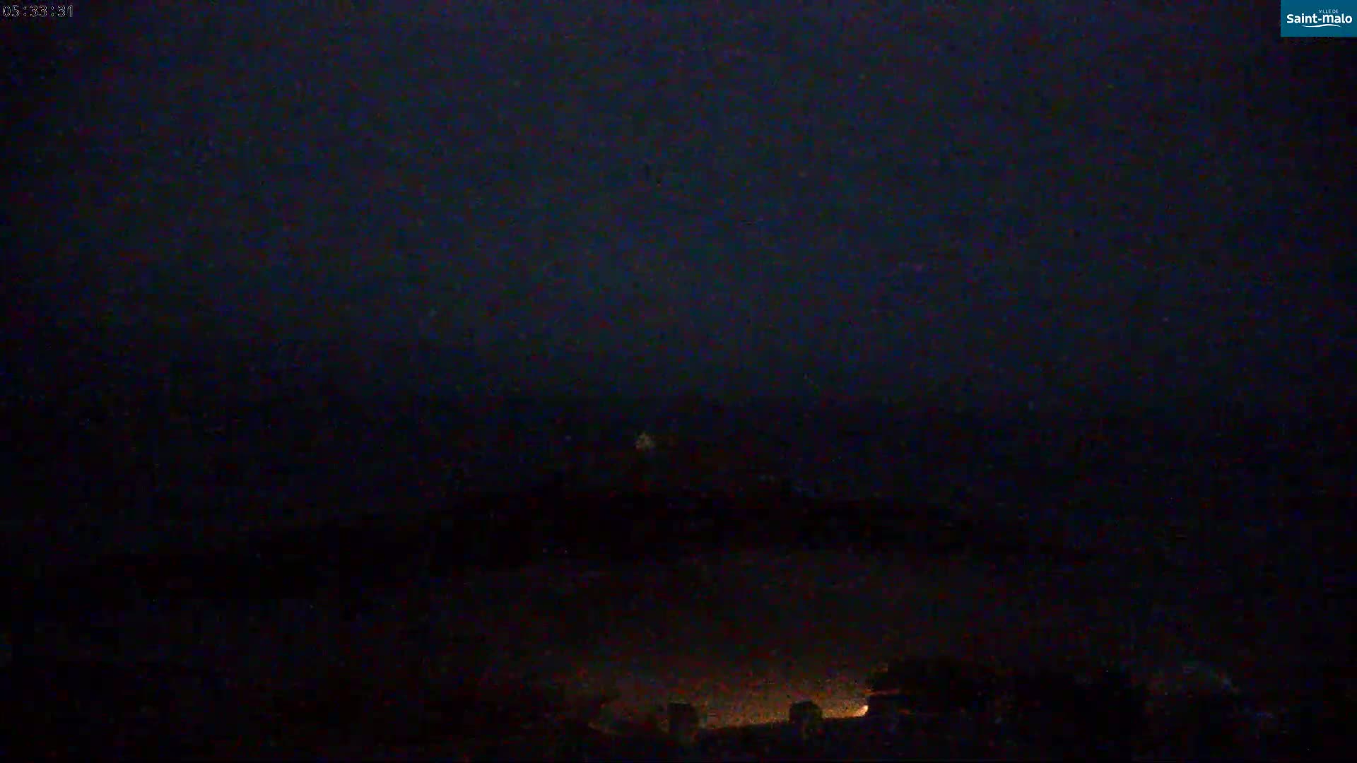 A dark, nighttime scene shows a dimly lit landscape with a few faint lights visible in the distance under a dark sky.