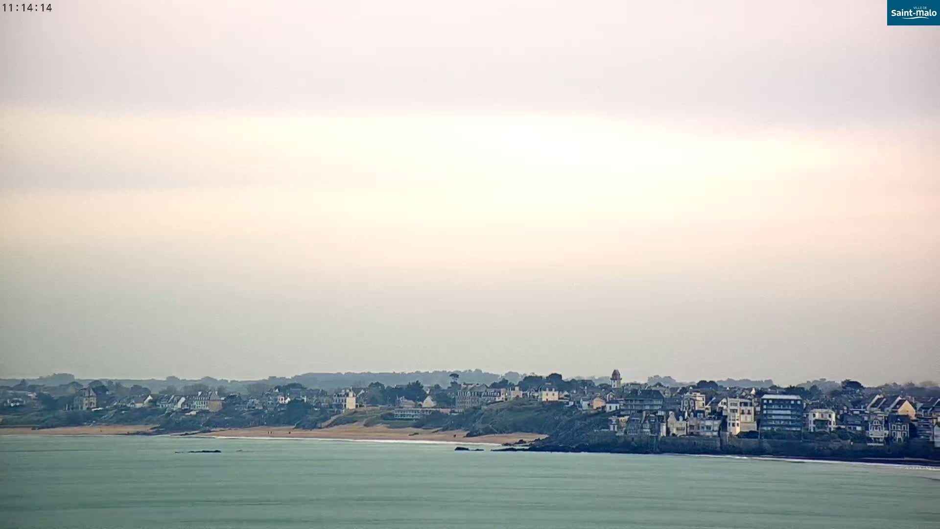 A coastal town sits on a gently sloping hill overlooking a calm, light-green ocean under a hazy, overcast sky.