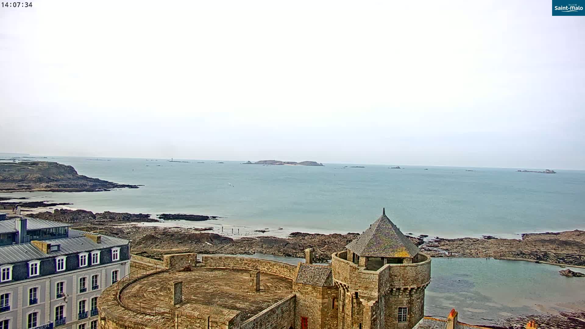 A stone castle turret overlooks a calm, light-blue ocean with rocky shores and distant islands under an overcast sky.