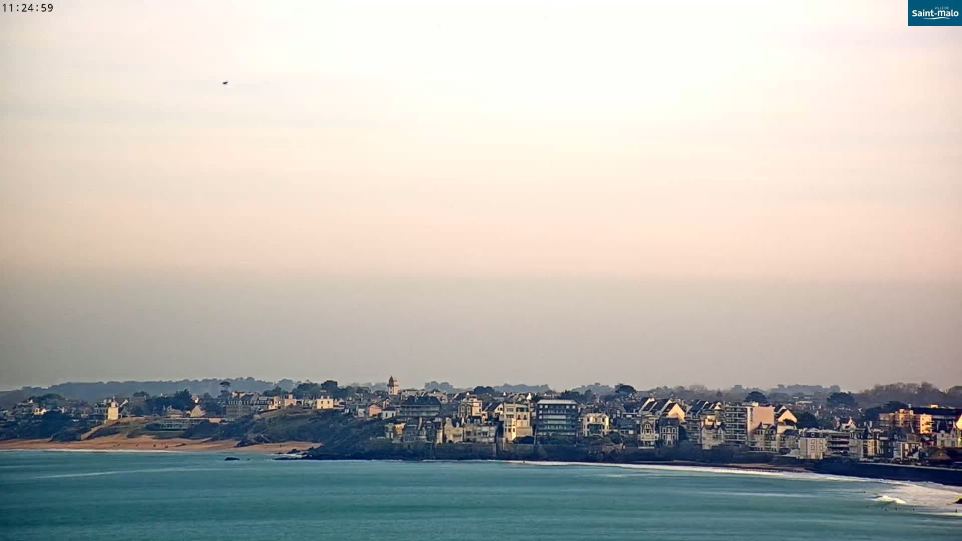 A coastal town with buildings lining a beach is visible across a calm ocean under a hazy sky.