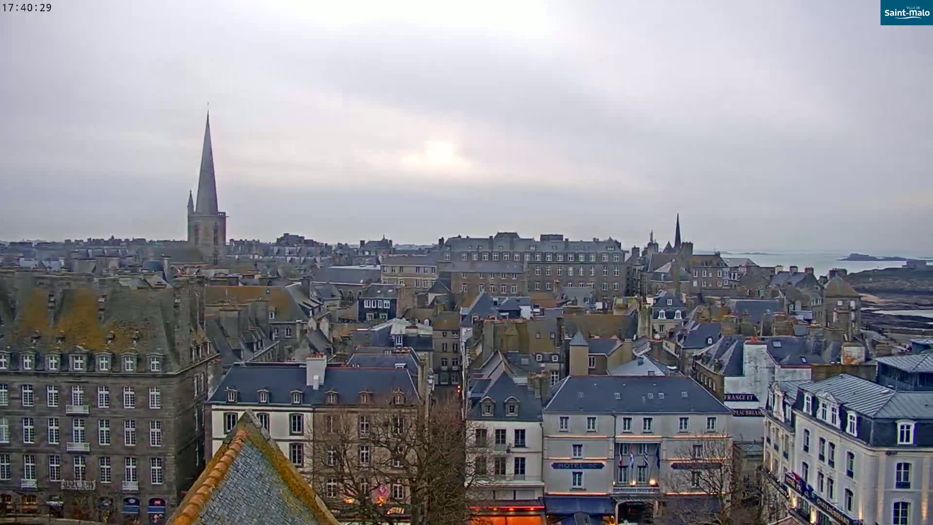A sprawling cityscape of grey-roofed buildings, punctuated by a tall church spire, stretches to the coast under an overcast sky.