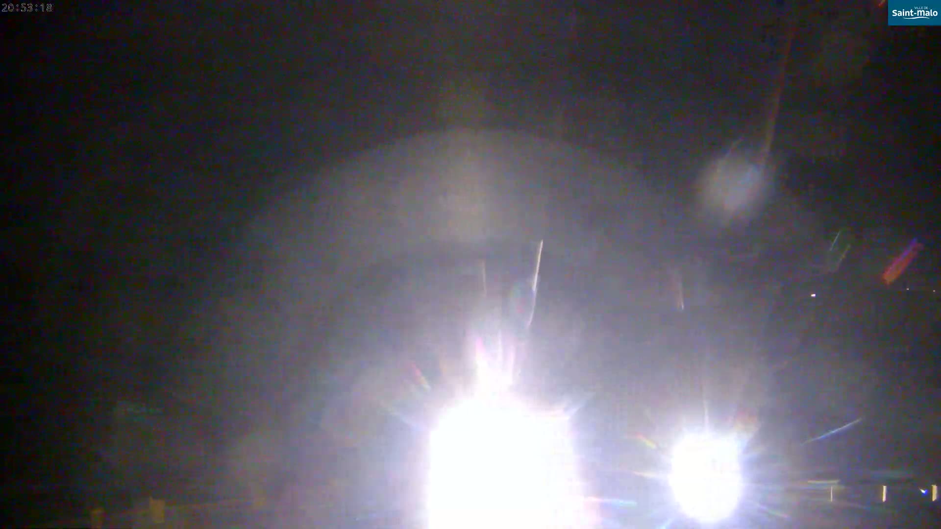 A nighttime outdoor scene shows bright headlights of a vehicle approaching, with lens flare obscuring the view.