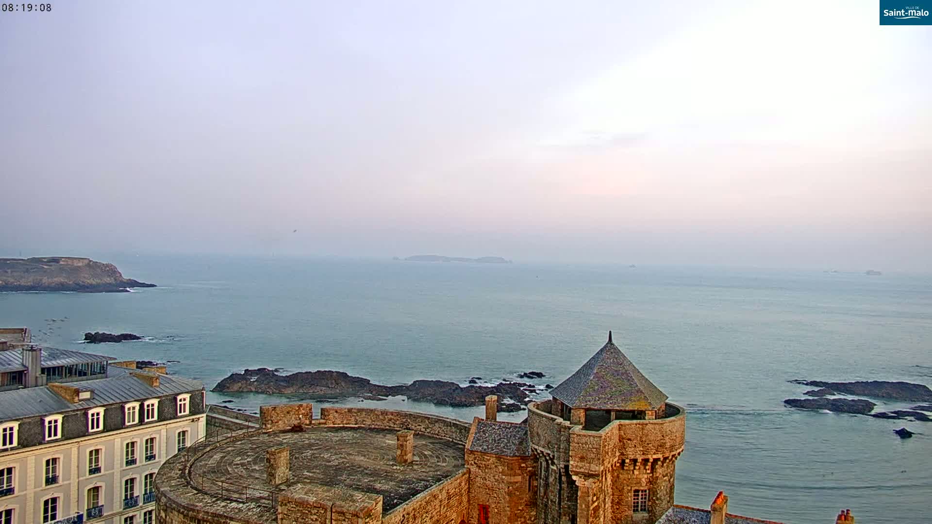 A stone castle overlooks a calm, pale-blue ocean under a hazy sky.