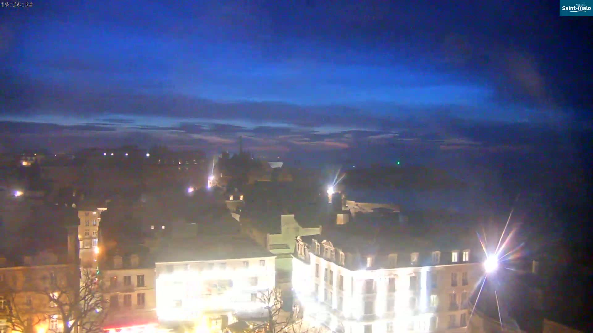 A nighttime view of a city with low-lying clouds under a twilight sky, showing illuminated buildings and streets.