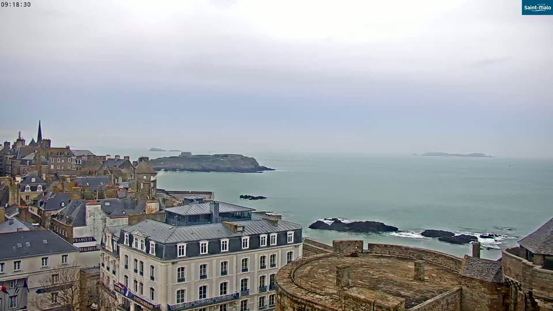 A coastal town with gray stone buildings overlooks a calm, gray-blue sea under an overcast sky.