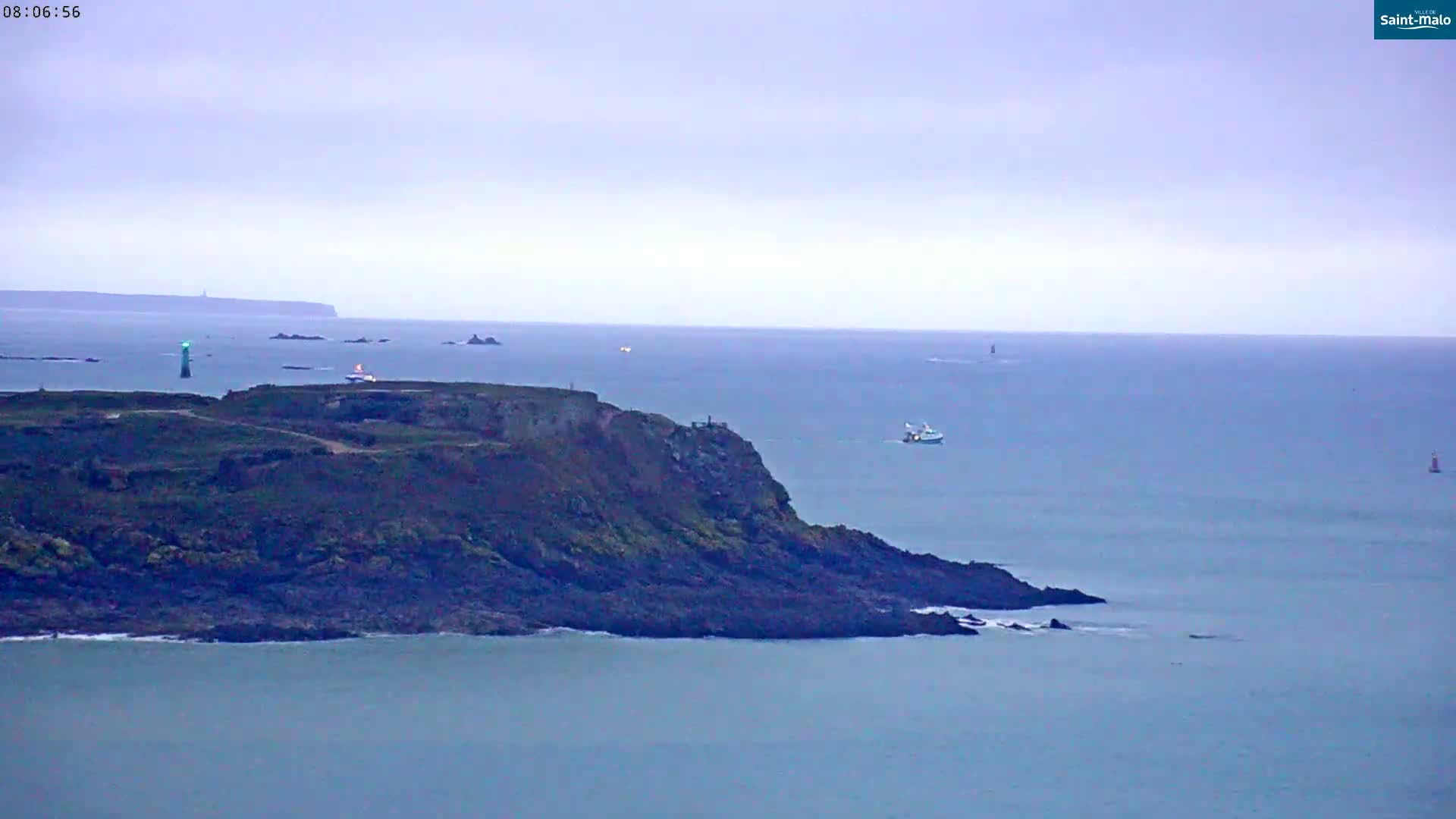 A rocky, dark-colored coastline extends into a calm, light-blue sea under an overcast sky, with several small boats and distant lighthouses visible.