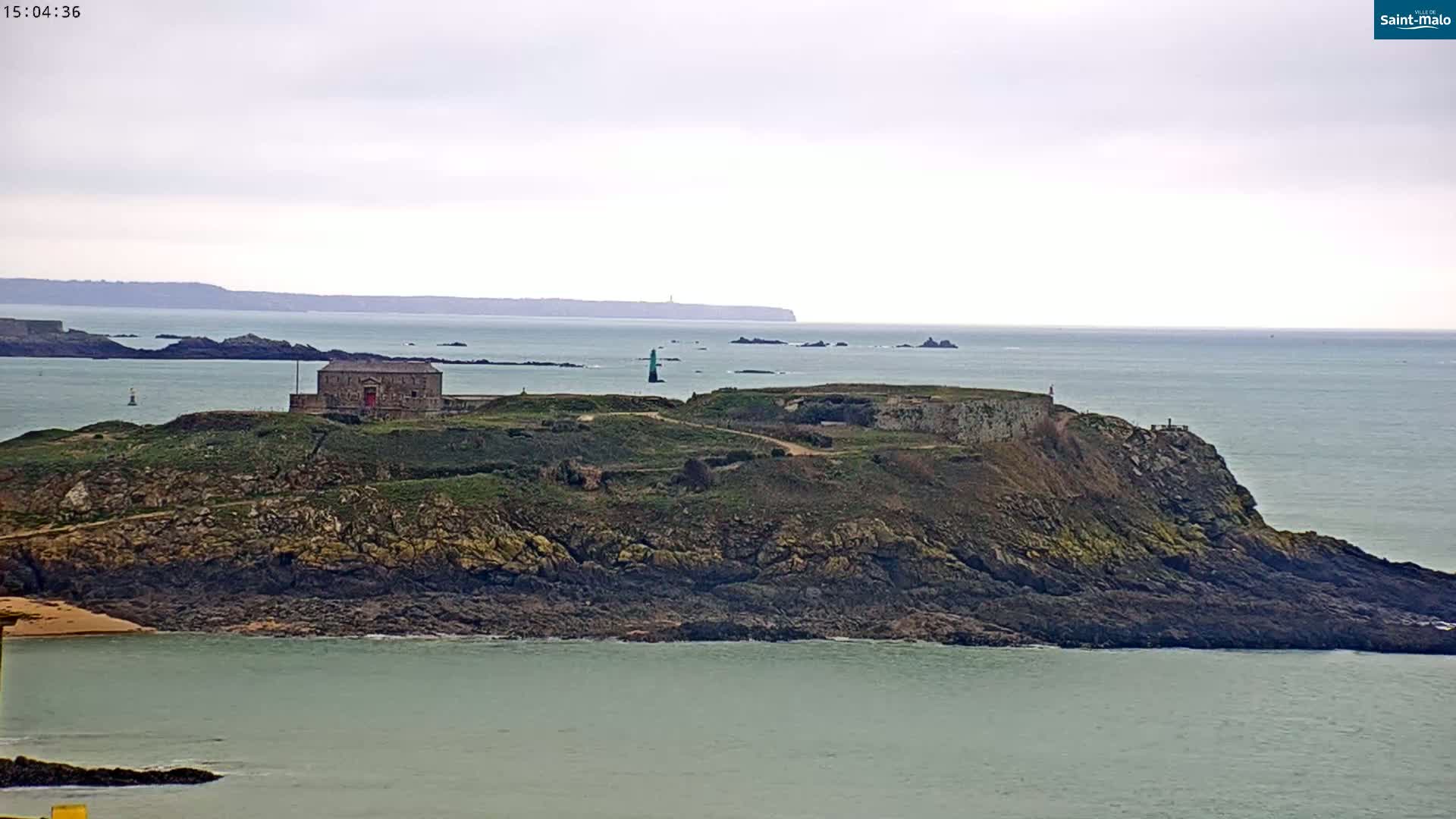 A rocky island with a stone building and a lighthouse, under a cloudy sky, is surrounded by calm sea water.
