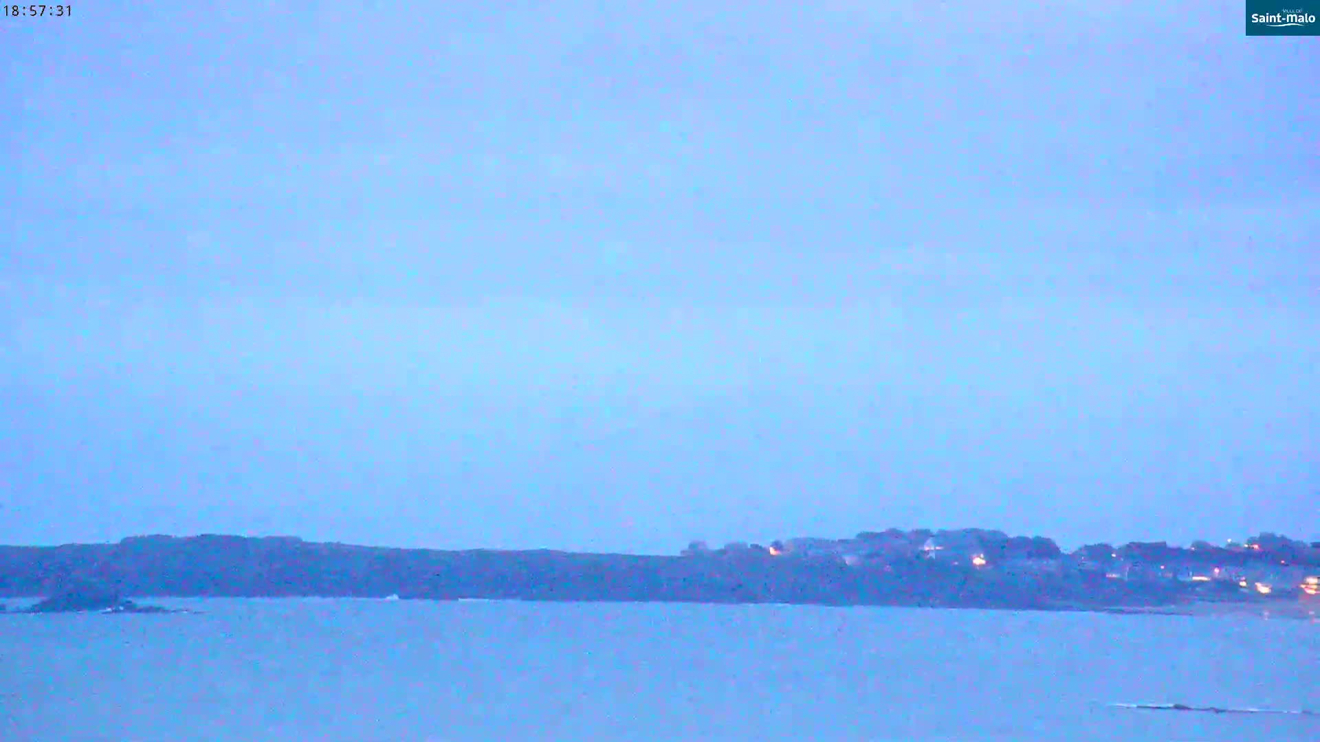 A calm body of water is shown with a dark landmass in the background under a pale blue, twilight sky.