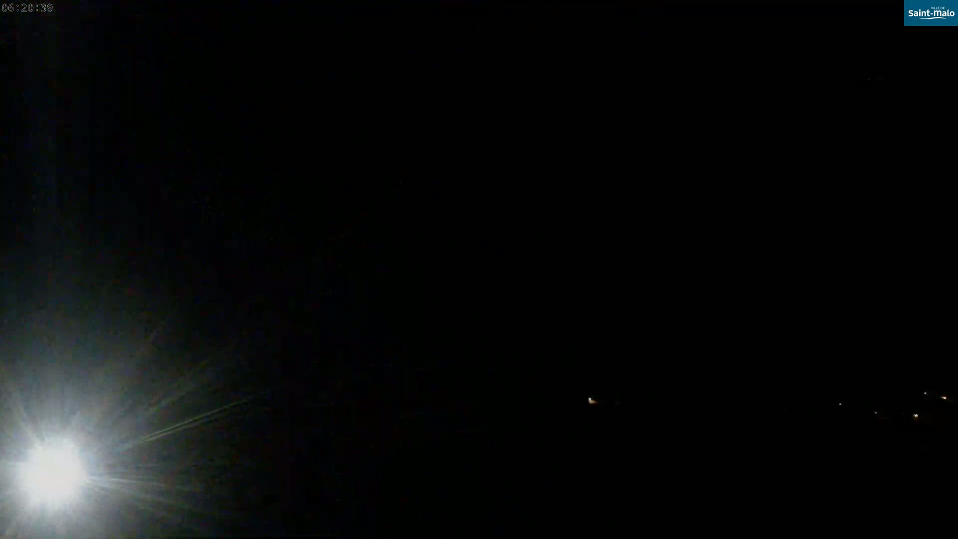 A bright light flares in the lower-left corner of a mostly dark, nighttime outdoor scene with a few distant lights visible on the horizon.