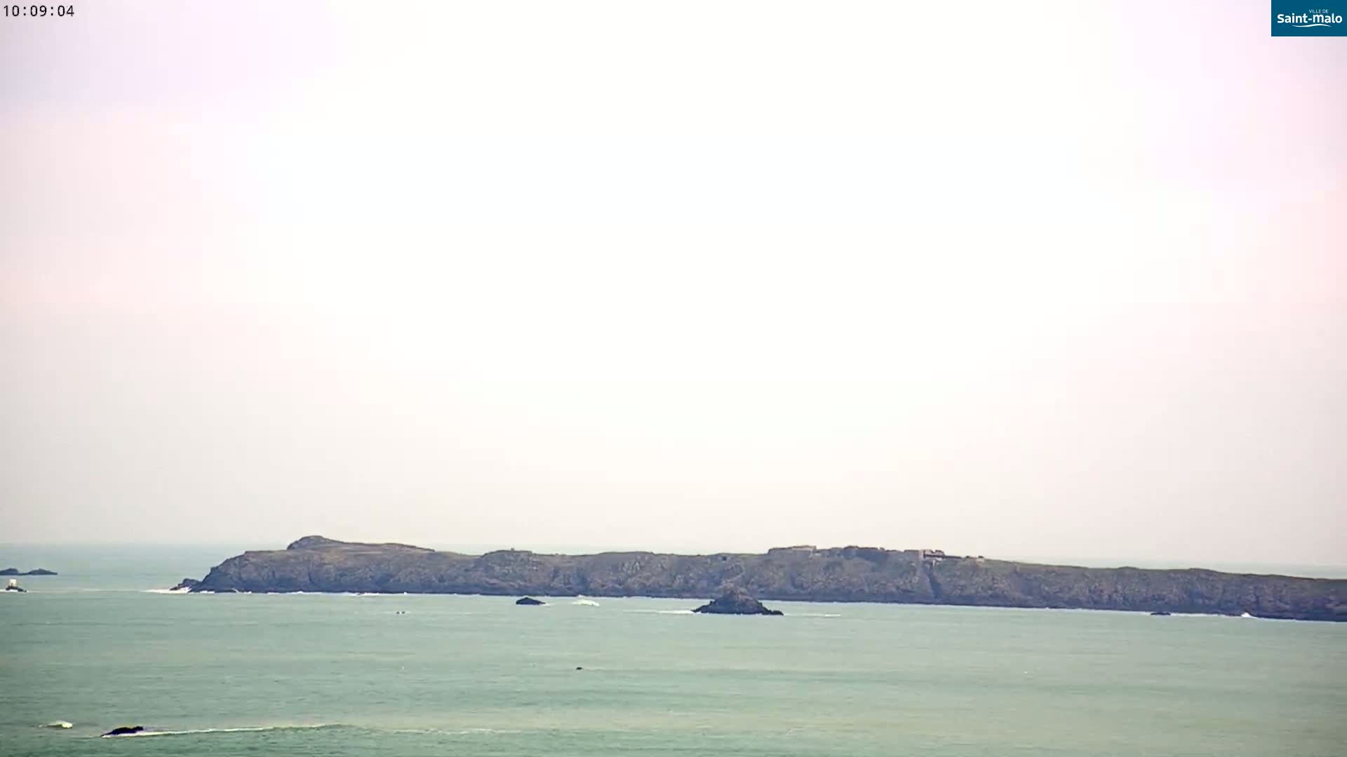 A long, dark island sits on a calm, light-green ocean under a hazy sky.