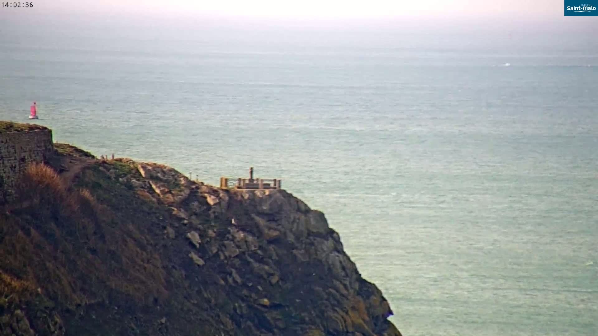 A rocky cliff overlooking a calm, light-green ocean with a small structure on its peak and a sailboat visible in the distance.
