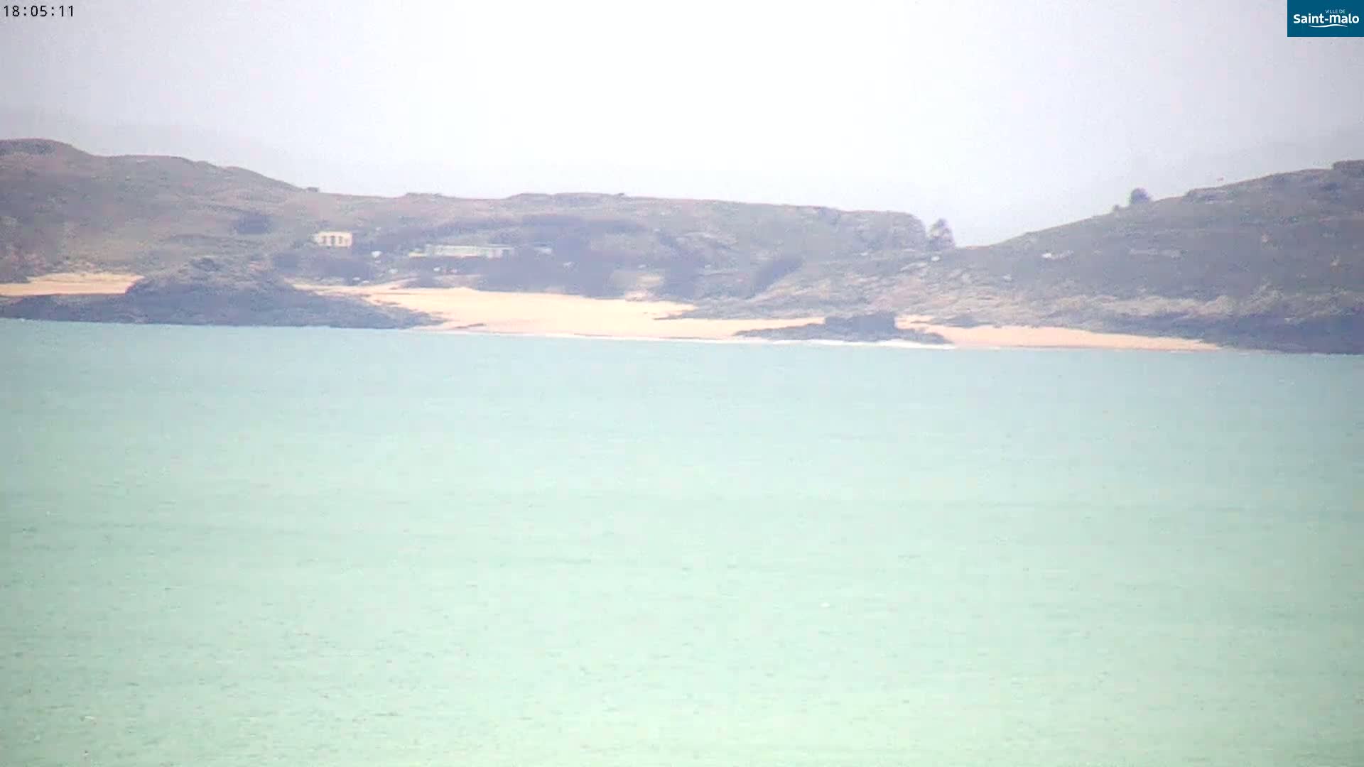 A calm, light-green ocean stretches out before a hazy coastline featuring dark-colored hills and a small, pale sand beach.