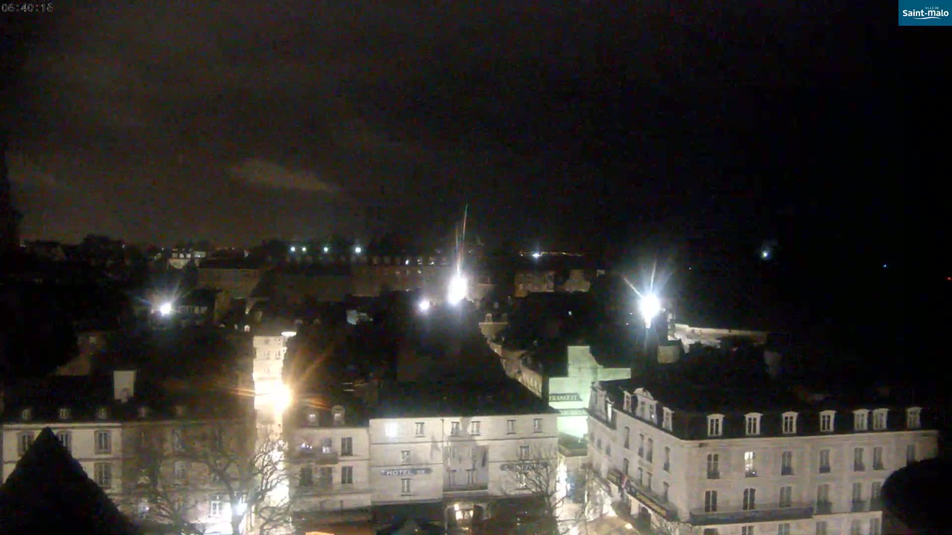 A nighttime, high-angle view of a city with buildings illuminated by streetlights under a dark, cloudy sky.
