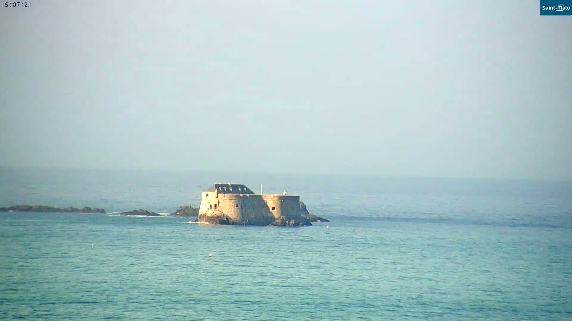 A stone fortress sits on a small island in a calm, hazy ocean.