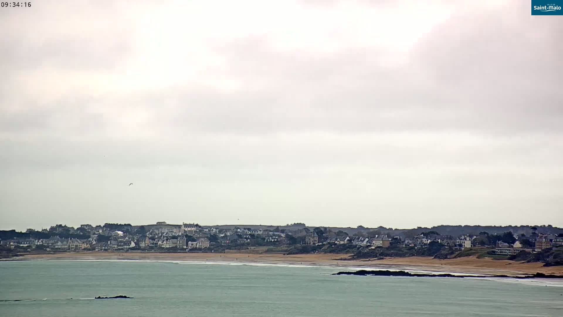 A coastal town with light-colored buildings stretches along a sandy beach under a mostly cloudy sky.