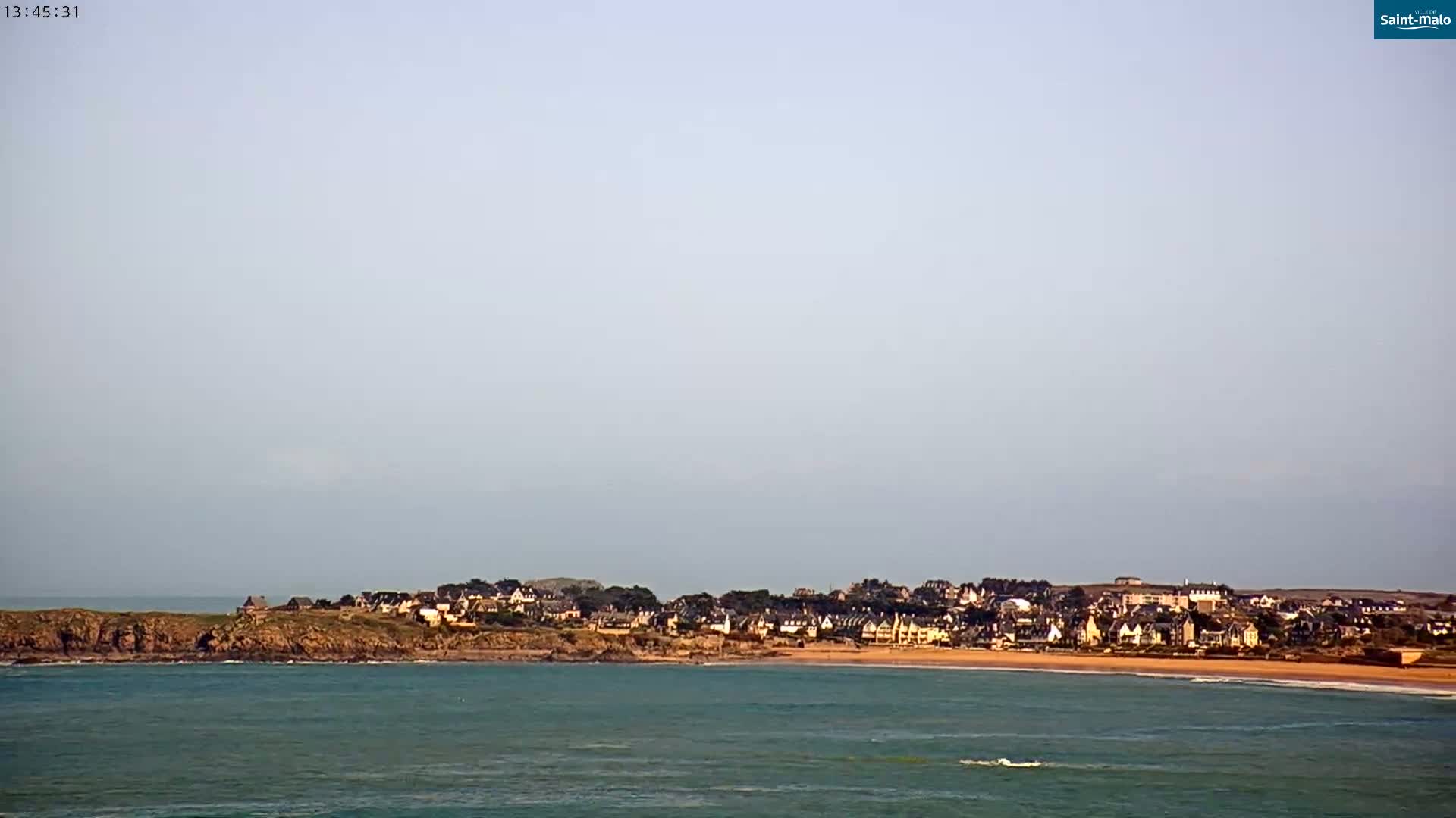 A coastal town sits on a rocky shoreline with a calm, light-blue ocean under a clear, light-grey sky.