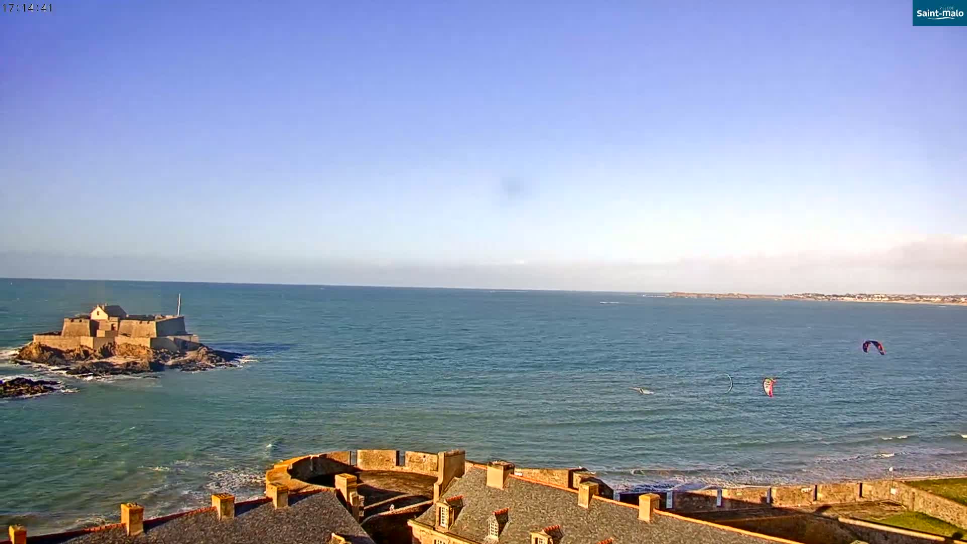 A coastal scene shows a stone fortress on an island in a calm ocean under a clear blue sky, with kitesurfers visible in the distance.