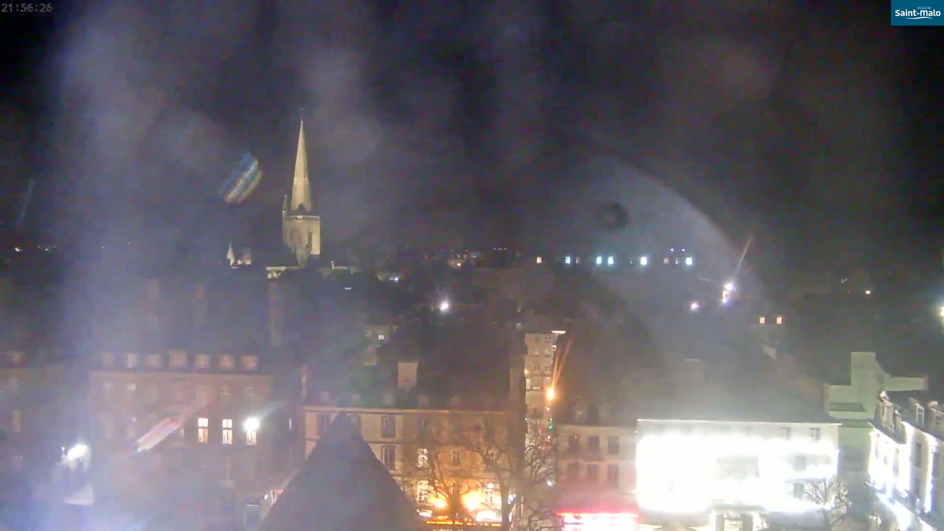 A nighttime view of a town, featuring a tall church spire and numerous buildings illuminated by artificial light, under a hazy sky.