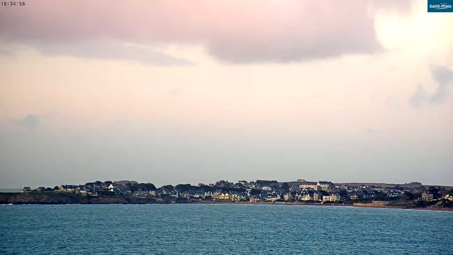 A coastal town sits on a low-lying peninsula under a mostly cloudy, pale pink and gray sky, with a calm sea in the foreground.