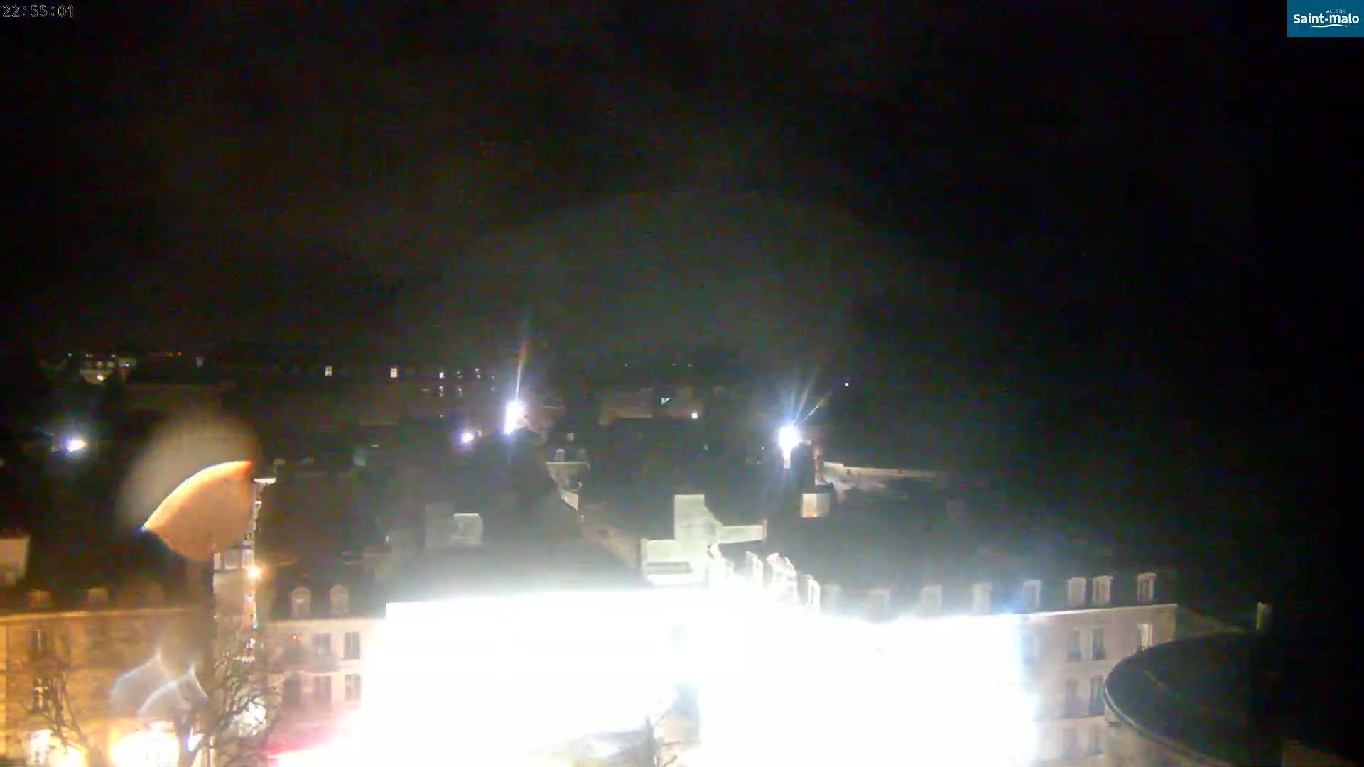 A nighttime, high-angle view of a town's rooftops and buildings, illuminated by artificial lights.