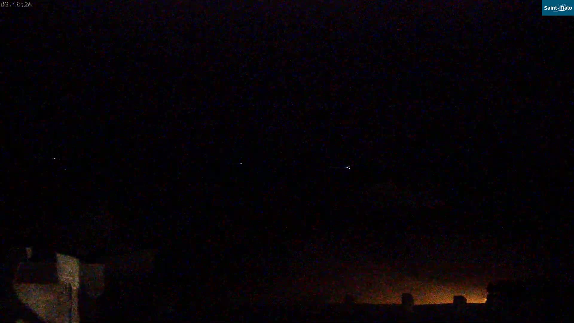 A dark nighttime scene shows a few faint lights in the sky above a dark, low horizon with some faint, distant lights.