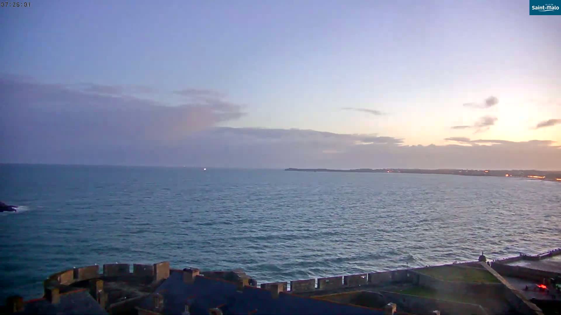 A calm ocean at sunset is viewed from a stone wall, with a faint landmass visible in the distance under a mostly clear sky.