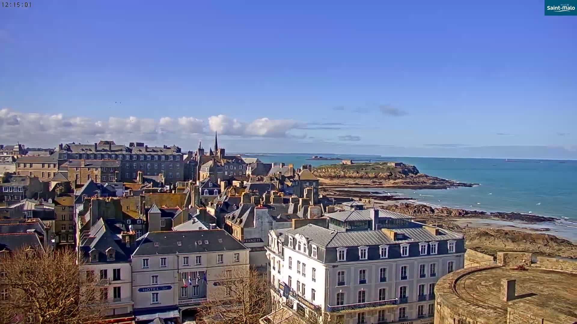 A coastal city with stone buildings and a church steeple overlooks a rocky shoreline and calm blue ocean under a mostly sunny sky.