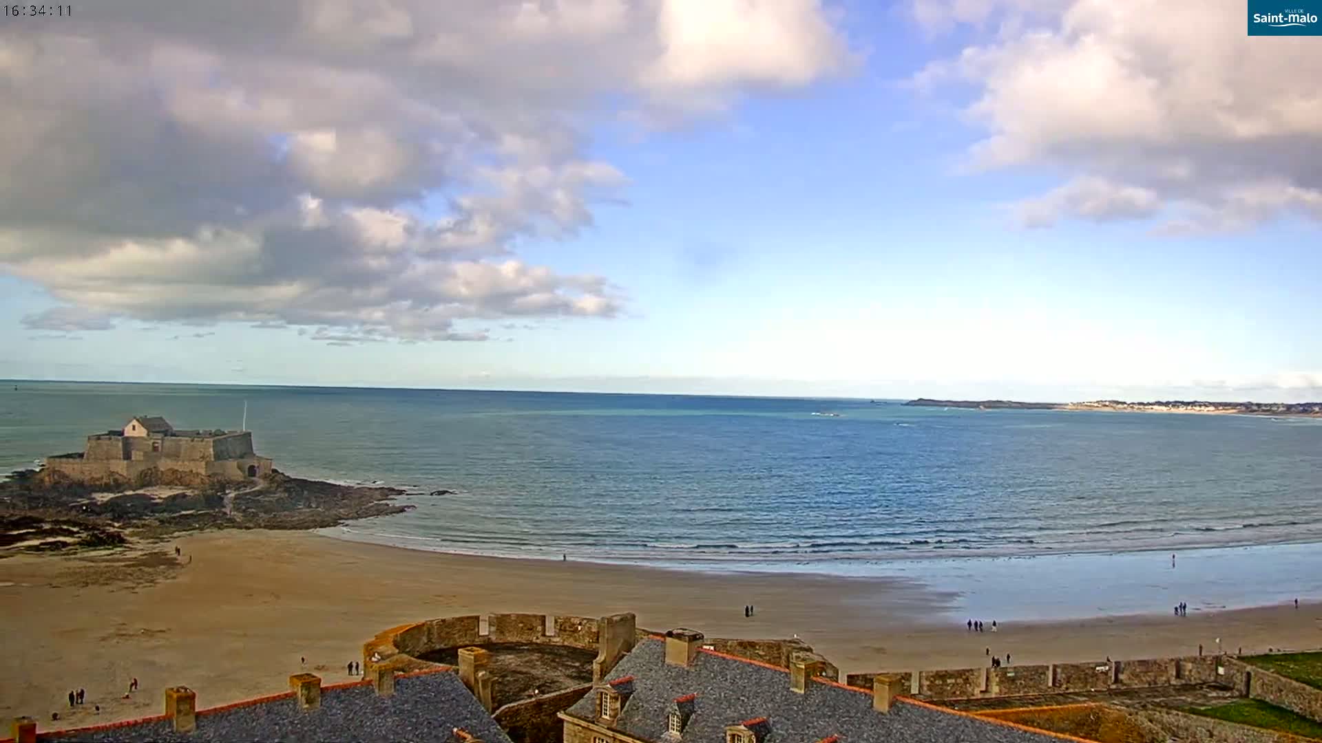 A sandy beach stretches out to a calm sea under a partly cloudy sky, with a stone fort and coastal town visible in the distance.