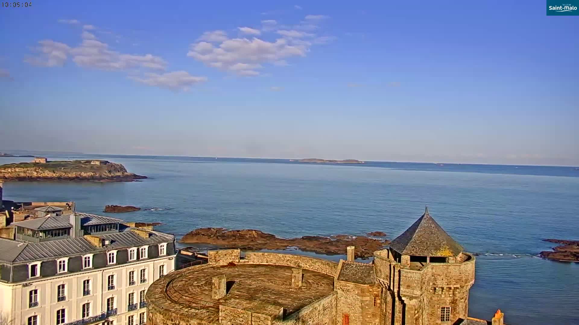 A coastal scene on a partly sunny day shows a stone castle overlooking a calm ocean with islands in the distance.