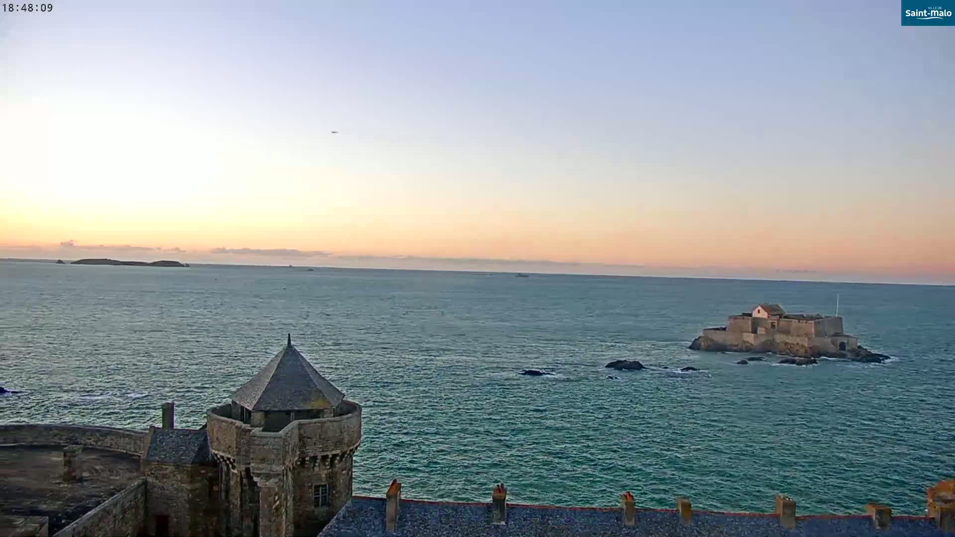 A stone tower overlooks a calm sea at sunset, with a small island fortress visible in the distance.