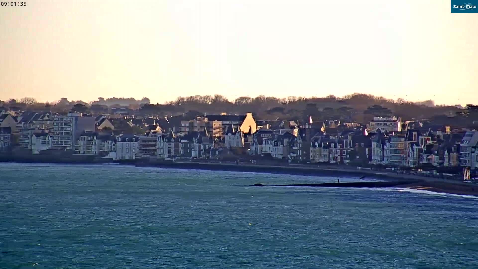 A seaside town with many closely-packed buildings is visible across a body of choppy water under a clear, light-colored sky.