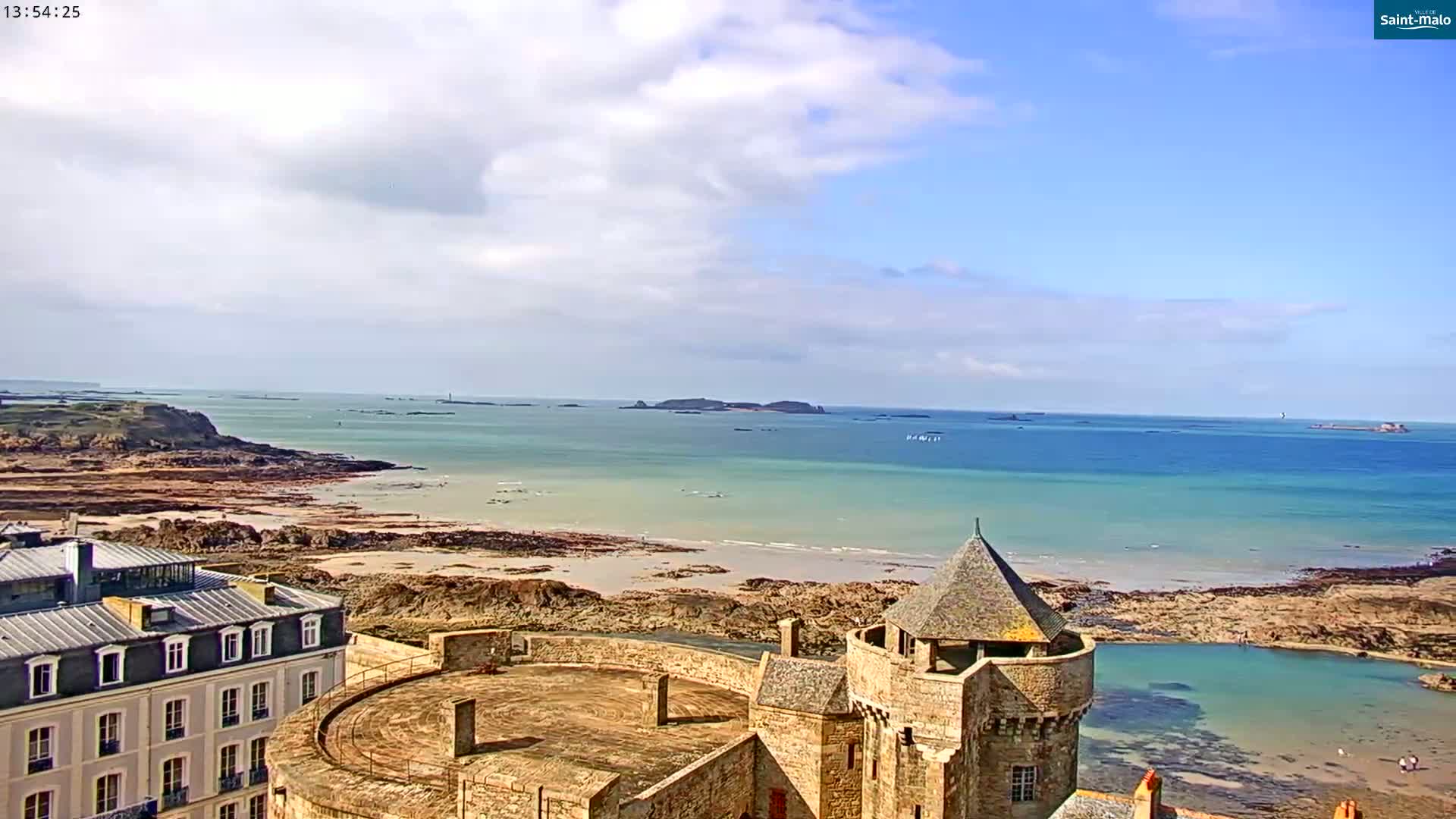 A partly cloudy sky overlooks a stone castle situated near a sandy beach and calm ocean with several small islands in the distance.