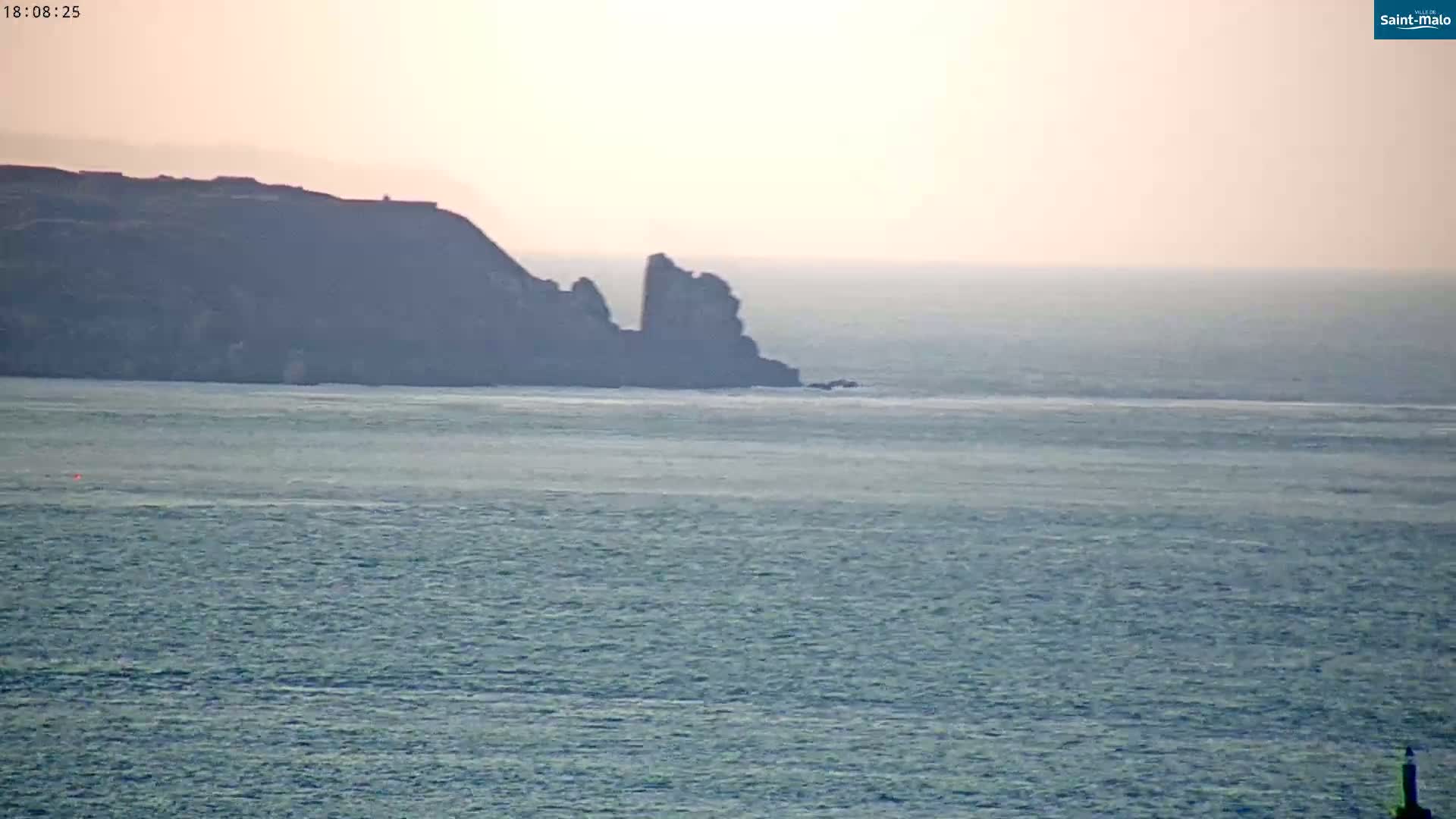 A hazy sunset illuminates a calm ocean with a dark, rocky headland in the distance.