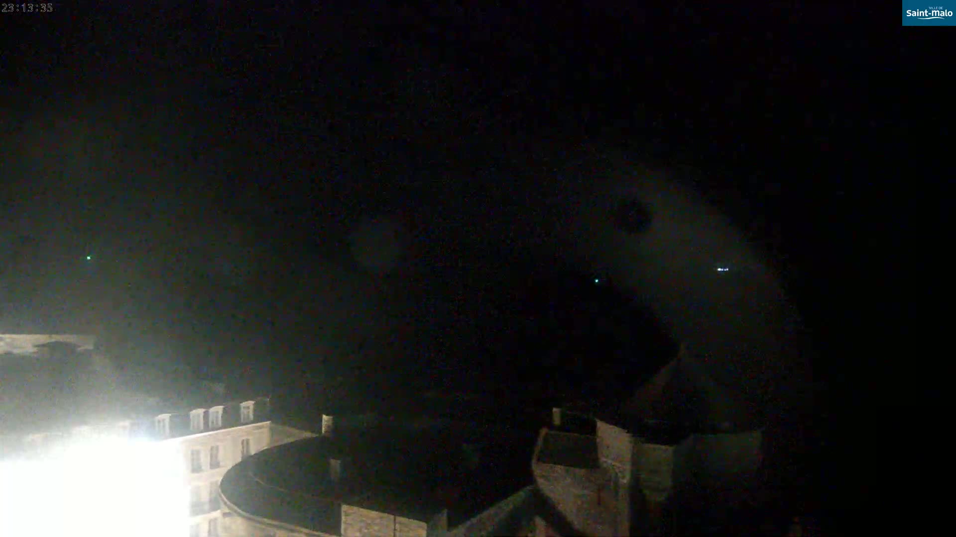 A nighttime view of a city, possibly coastal, with a dark, cloudy sky showing faint lights in the distance.