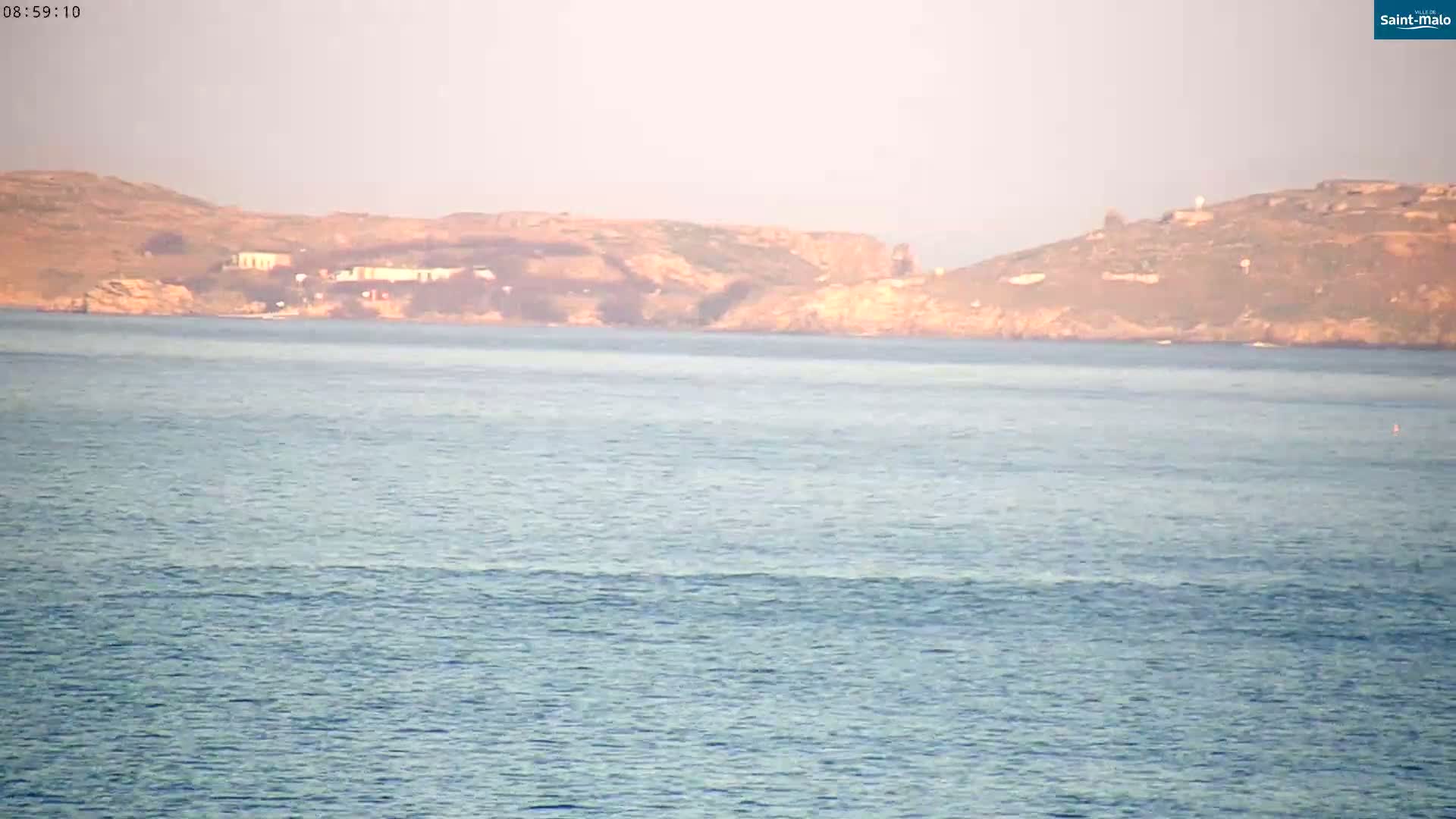 A calm body of water shows gentle waves lapping against the shore, with a distant landmass under a hazy sky.
