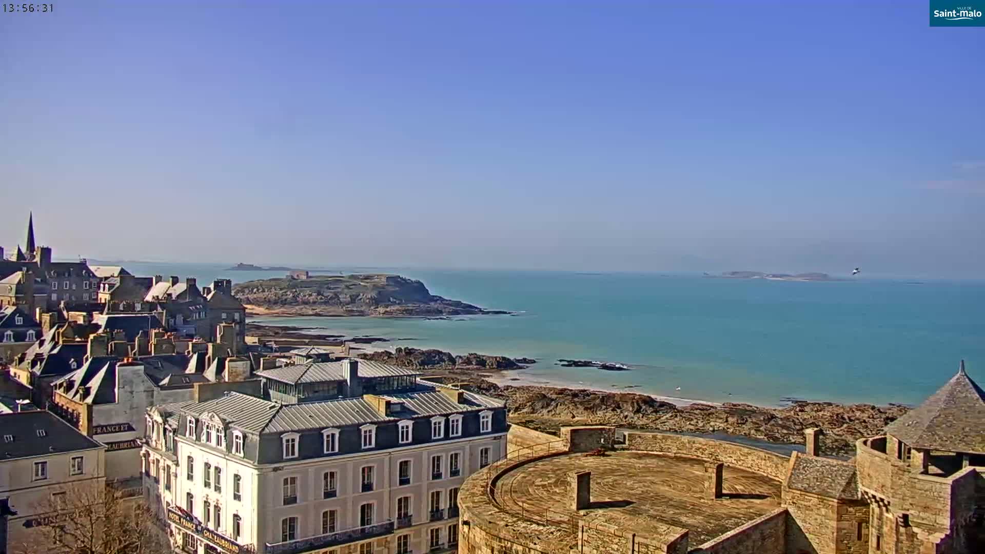An elevated view shows a coastal town with stone buildings, a partially visible castle, and a clear blue ocean under a bright sunny sky.