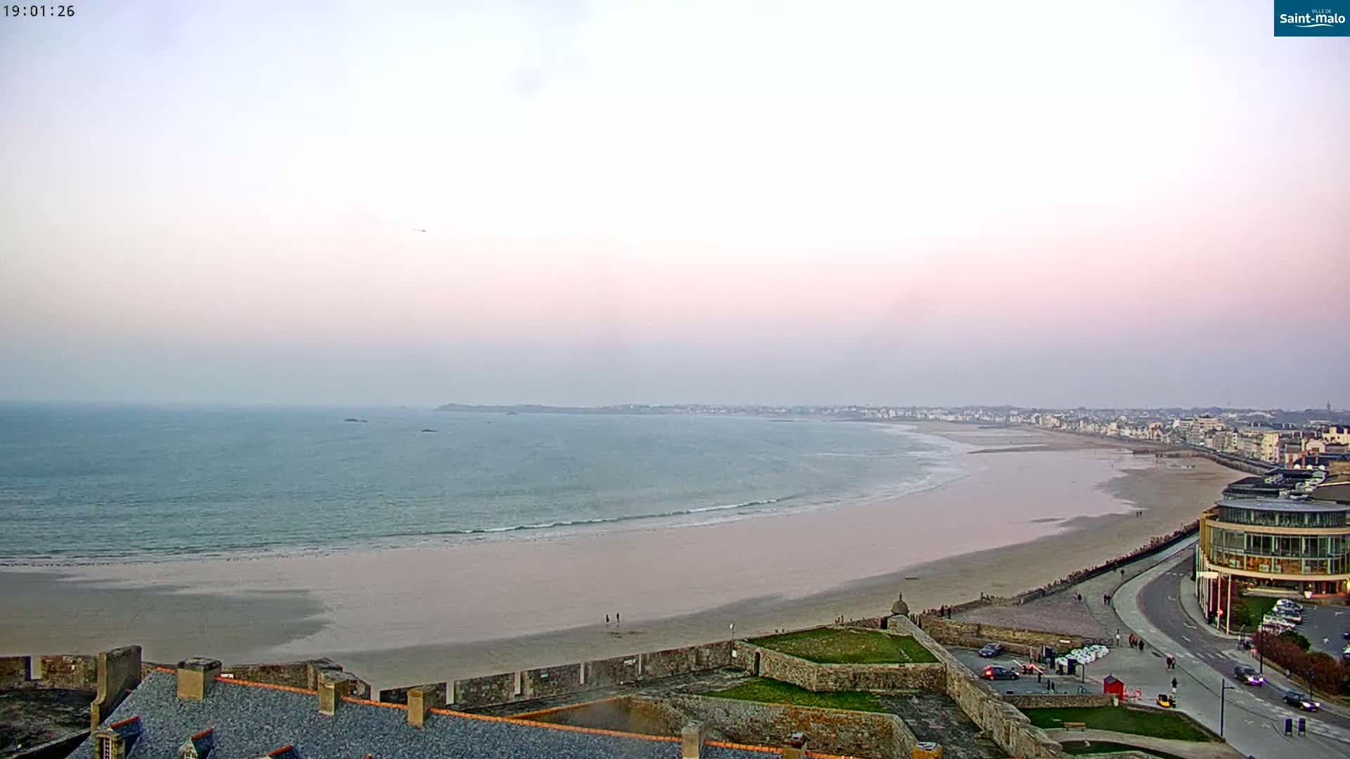 A long, sandy beach curves along a calm ocean under a pale pink twilight sky.
