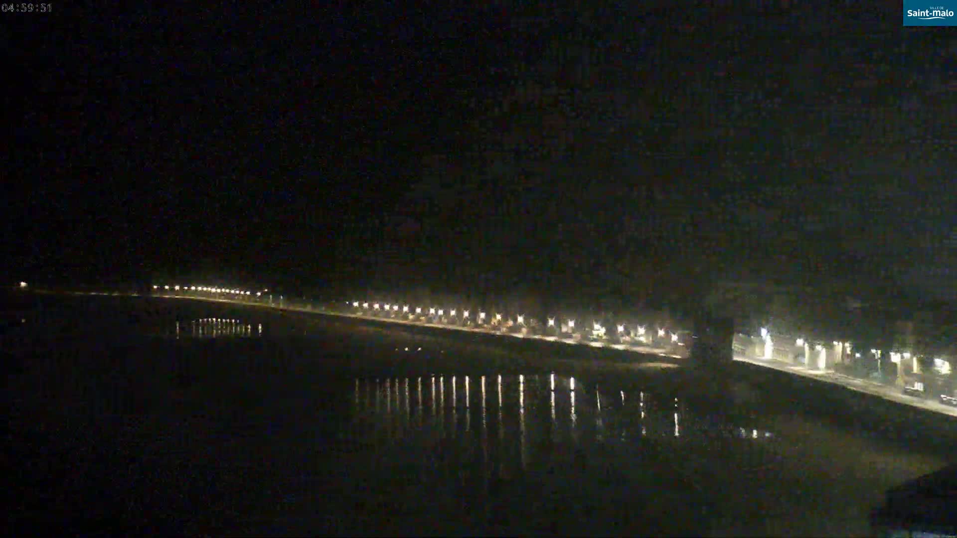 A nighttime view of a waterfront road with lights reflecting in calm water under a dark sky.