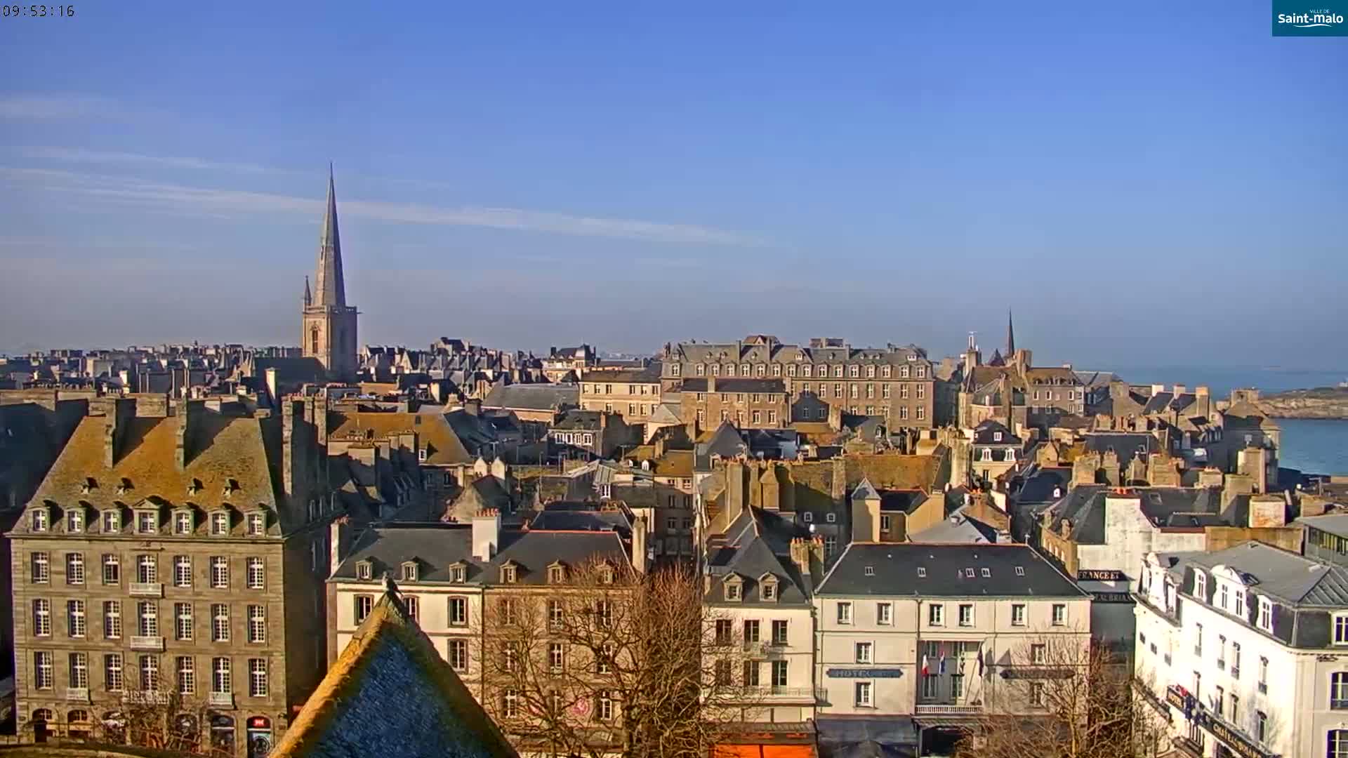 A panoramic view of a city with many buildings under a clear, sunny sky shows a body of water in the distance.