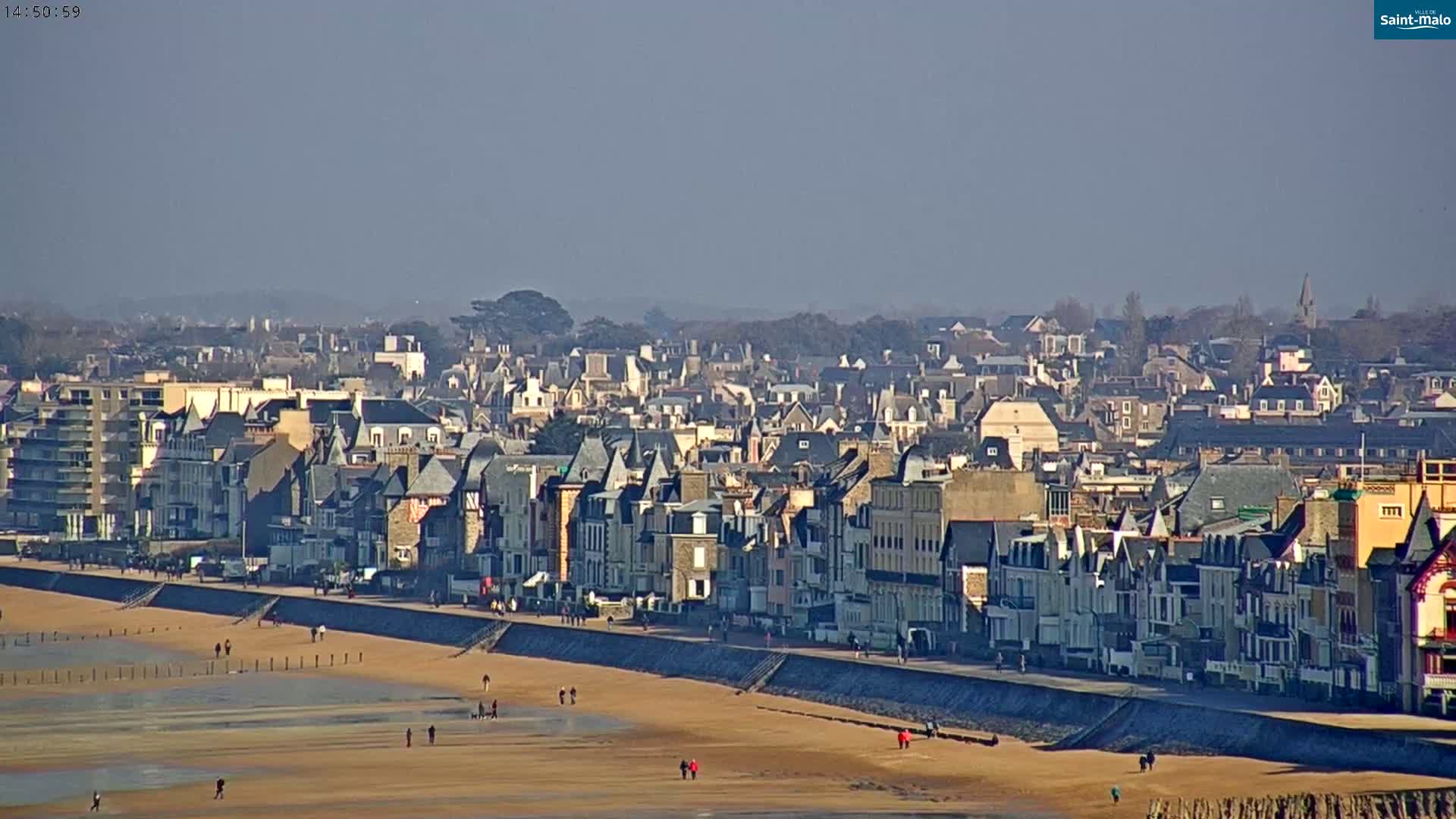 A sandy beach with a few people walking on it stretches out in front of a long row of closely-packed buildings along a seawall, with more buildings extending into the hazy distance under a clear, sunny sky.
