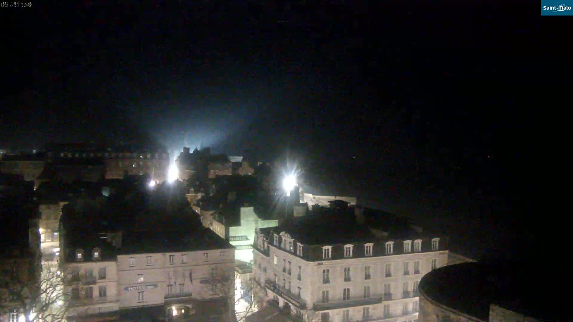 A nighttime, high-angle view of a city's buildings illuminated by streetlights under a dark, possibly foggy sky.